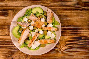 Fresh salad with chicken meat, feta cheese, avocado, green olives and lettuce leaves in ceramic plate on wooden table. Top view
