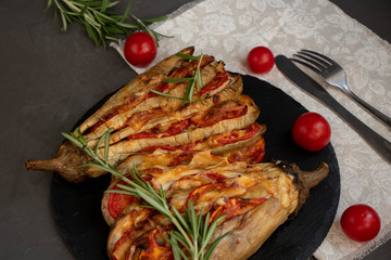 Dietary fan of eggplant white filled with tomatoes and cheese. Nearby are cherry tomatoes, rosemary branches. It is worth soy sauce and metal appliances. On a dark background.