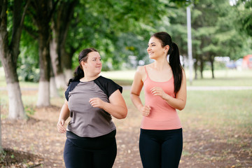 Fototapeta premium Friendship, personal trainer, group workout, weight loss, sports and health care. Young fit woman support and motivate her overweight female friend at outdoor jogging