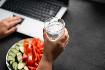 Healthy snack at workplace. Woman working at computer, drinking water and snacking with sliced vegetables. Low calorie food, dieting, lifestyle and weight loss