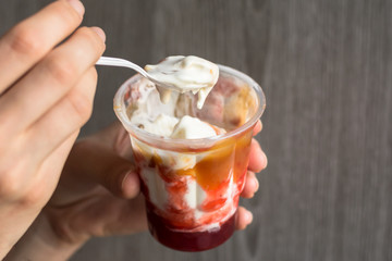 ice cream in a plastic cup. Below is strawberry jam. Above the liquid caramel. Above the glass is a plastic spoon with spreading caramel. The cup is in the human palm
