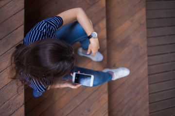 Brunette girl with a large wrist watch, sits on wooden steps and checks a smartphone. The view is clearly from above.