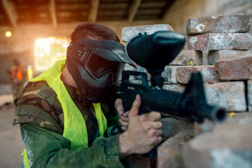 Male paintball player with protective mask and uniform aiming with paintball gun 