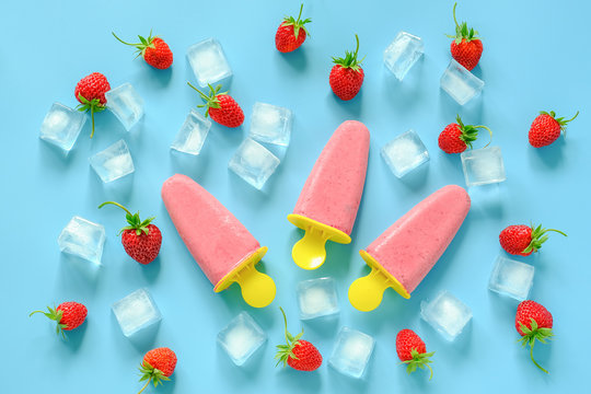 Homemade popsicles. Natural ice cream in bright plastic molds, strawberries and ice cubes on blue background. Top view Flat lay