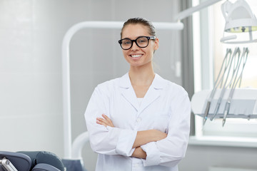 Waist up portrait of young female doctor wearing glasses smiling at camera while posing in office, copy space