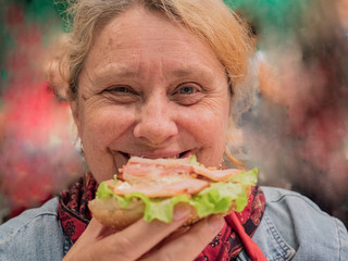 Retired elderly woman with red hair is tasting large hamburger cooked in high-tech kitchen. Master class on cooking meat dishes and hamburgers