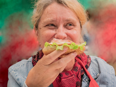 Retired elderly woman with red hair is tasting large hamburger cooked in high-tech kitchen. Master class on cooking meat dishes and hamburgers - Powered by Adobe