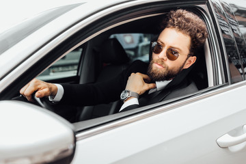Portrait of a stylish bearded man in sunglasses behind the wheel of a white car, an expensive watch...