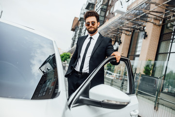 A handsome successful businessman in full suit and sunglasses opens his car on the streets of the city near the modern office center.