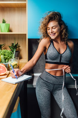 Woman fitness dietolog at home kitcvhen. Beautiful girl in sportswear measuringand write recipe...
