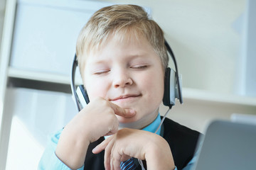 Cute little boy in suit listening to music or audio tutorial on headphones at the office background.