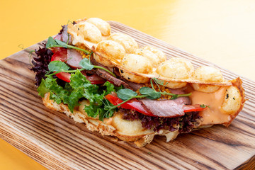 Bubble waffle sandwich with ham, tomato and lettuce. On a wooden board.