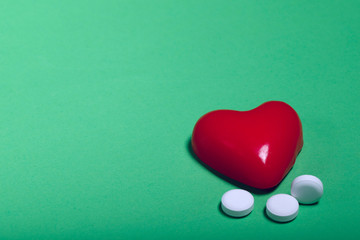 Fototapeta premium A group of white medicine pills and a red heart shape on a green background. Cropped shot, horizontal, top view. The concept of medicine and health.