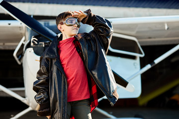 Fototapeta na wymiar close up view of a boy in aviator glasses watching aviation show in the sky, dressed in large black leather pilot jacket, red shirt and jeans, white propeller plane stands behind him. Outdoor shot.