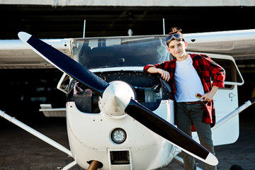 cute boy in aviator glasses, red plaid shirt, holds screwdriver and stands near white light private propeller air jet that is parked outside hangar, looks in camera with friendly smile.