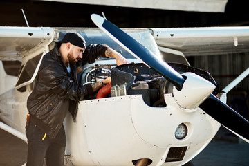 handsome young pilot in black leather jacket and instruments in his jeans pockets, checking up thoroughly motor compartments of his light propeller air jet outside hangar building. © alfa27