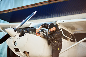 young mechanic engineer inspecting light single-engine propeller airplane constructions, fixing tube, checking engine, working hard all day long outside aircraft shed.