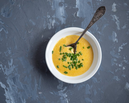 Soup of mashed potatoes with vegetables and cheese sprinkled with green onions in a deep white plate, a tablespoon on a gray background, top view