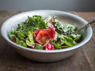 Fresh salad from tomatoes, cucumbers and aromatic herbs, dressed with sour cream in a deep aluminum bowl with a spoon, on an old wooden table, shot from close range