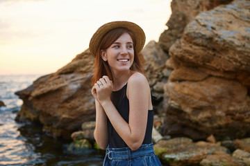Portrait of young positive ginger freckles lady in hat, broadly smiles and enjoy the morning on the seaside, looks cheerful and happy.