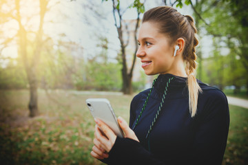 Young smiling girl making sport and running in the park using her phone to listen the music with wireless headphones on sunset in the city watching the screen and having communication talking chat