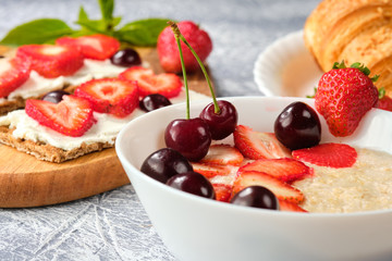 white bowl of porridge with strawberries and cherries, croissants and crispbread with cheese and strawberries on a gray background