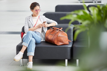 Pensive young girl in pink sunglasses sitting on leather sofa in airport and using gadget while...