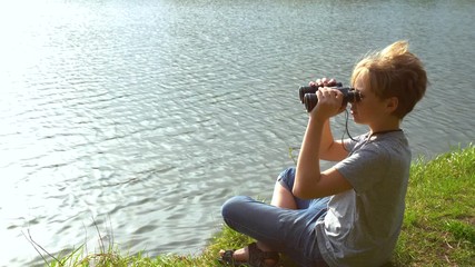 White kid sitting at seashore of river and looking through old black binoculars at something interesting in distance. Real time 4k video footage.