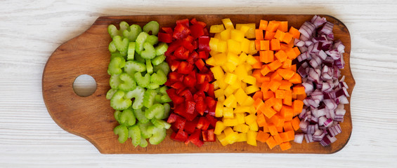 Overhead view, chopped fresh vegetables (carrot, celery, red onion, colored peppers) arranged on cutting board on a white wooden surface. Flat lay, from above, top view.