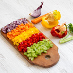 Chopped fresh vegetables (carrot, celery, onion, colored peppers) arranged on a cutting board on a white wooden background, side view.