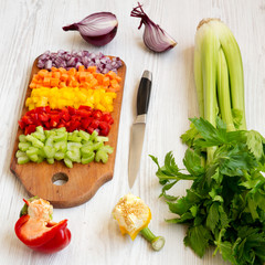 Chopped fresh vegetables (carrot, celery, onion, colored peppers) arranged on a cutting board on a white wooden surface, side view. Close-up.