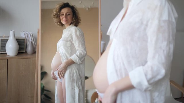 Happy Young Pregnant Woman Looking In Mirror On Her Belly. Motherhood, Pregnancy Concept.