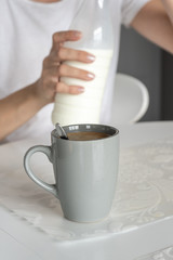 In the kitchen there is a gray cup with freshly prepared morning coffee on a white table. A woman close a bottle of milk after preparation. Vertical