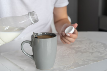 Close-up. There is a gray cup with freshly made morning coffee on a white table in the kitchen . A woman begin to pour milk from a bottle into a cup. 
