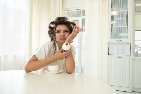 beautiful young brunette woman in hair curlers holding cupcakes in both hands sitting with cup of coffee in bright kitchen looking upset