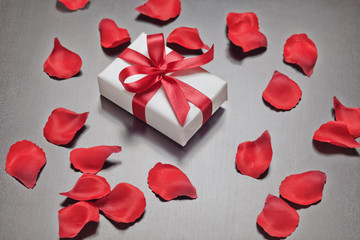White gift box with red ribbon and red rose petals on gray background