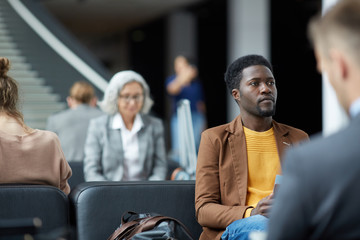 Serious young black man with beard sitting on seat with bag in airport and waiting for boarding