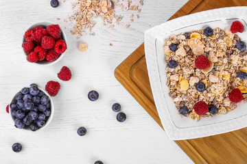 bowl of oat granola, muesli with fresh raspberries, blueberries on white wooden board for healthy breakfast. top view