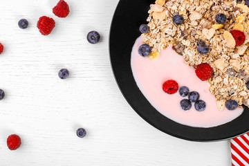 black bowl of oat granola, muesli with yogurt, fresh raspberries, blueberries on white wooden board for healthy breakfast. top view