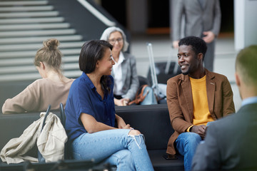 Positive handsome young Afro-American man with beard sitting on sofa and talking to lady in airport...