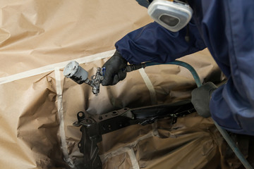 A male worker in jumpsuit and gloves paints with a spray gun a front frame part of the car body in black after being damaged at an accident. Auto service industry professions