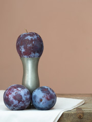 Three dark red, purple plums one on vase pedestal.
