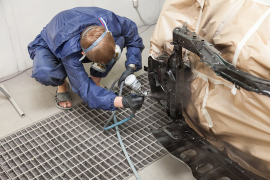 A male worker in jumpsuit and gloves paints with a spray gun a front frame part of the car body in black after being damaged at an accident. Auto service industry professions