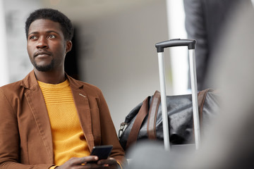 Serious handsome young Black man with beard sitting with luggage in airport and waiting for his...