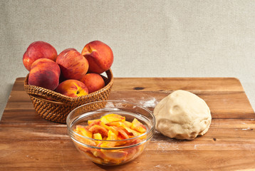 Top, front view, medium distance of ingredients to bake a peach pie, ball of dough, glass bowl of sliced ripe peaches and a basket of local, ripe peaches on a wood cutting board 