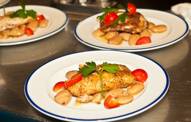 Top, front view, close distance of a freshly prepared red snapper filet on a bed of white bean salad, cherry tomatoes, parsley, on  round white plate, blue rim, at a wine dinner, tropical resturant