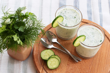 On a table on a wooden stand are two glass cups with a smoothie with cucumber and greens, a bunch of greens, metal spoons. Close-up.
