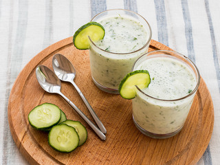 On a table on a wooden stand are two glass cups with a smoothie with cucumber and greens and metal spoons lie. Close-up.