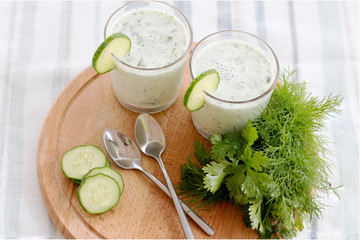 On a table on a wooden stand are two glass cups with a smoothie with cucumber and greens, a bunch of greens, metal spoons. Close-up.
