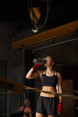 Dramatic portrait of tough sportswoman drinking water standing in boxing ring, copy space